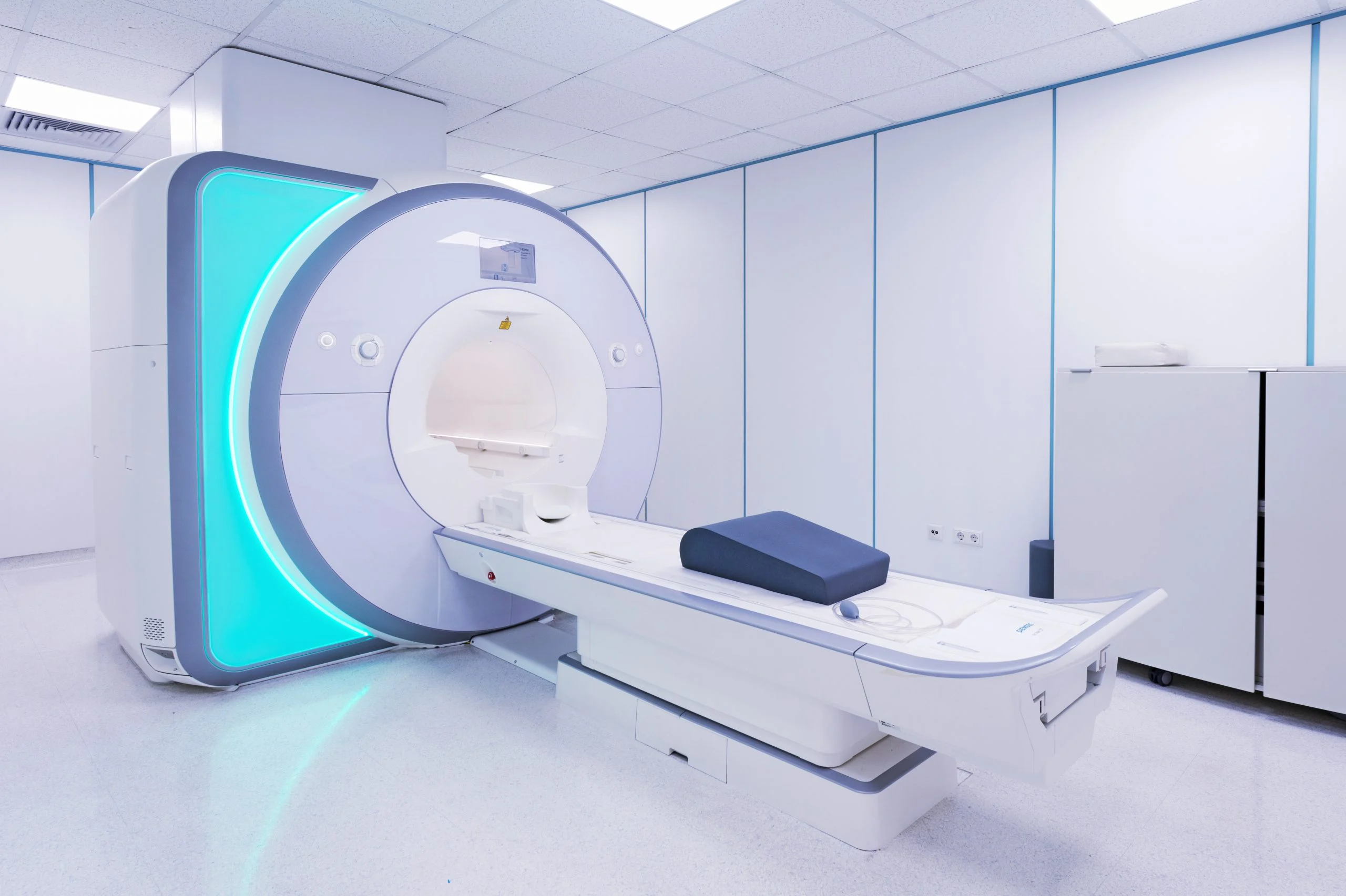 Modern MRI scanner with glowing cyan ring in a clean, white medical facility room with control equipment